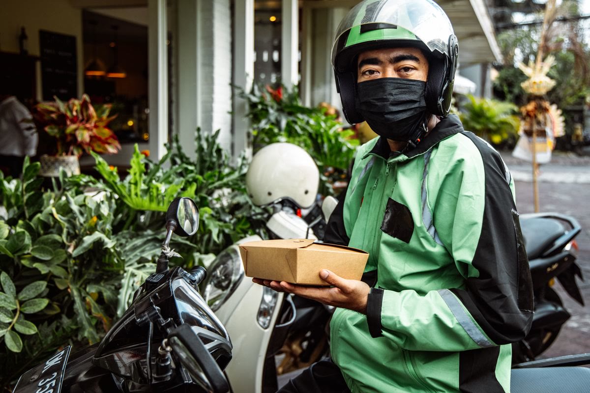 A delivery driver on a motorcycle wearing a green jacket and helmet, holding a food delivery box.